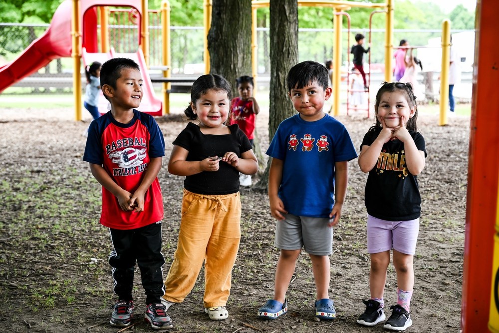 photo of 4 young children under the age of 5 who are standing amidst a playground of peers. They're all looking and smiling