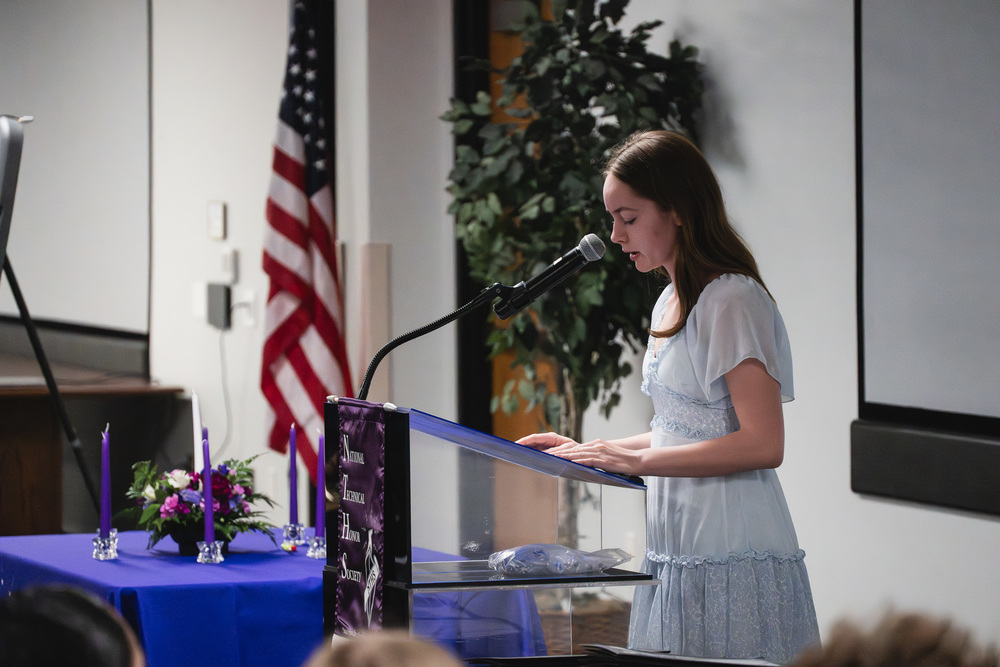A Van Buren Tech student in a light blue dress stands at a podium, speaking into a microphone during an indoor ceremony. An American flag and a large plant are visible behind her, and a table with purple candles and a floral arrangement sits nearby.