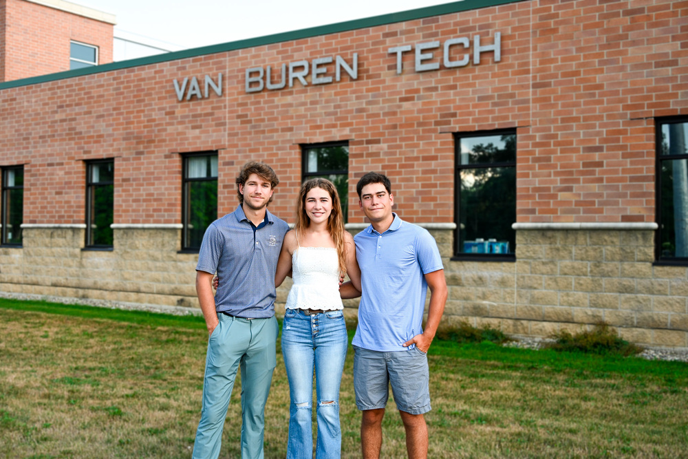 hree Sylvester siblings stand outside Van Buren Tech, smiling at the camera. The two brothers wear collared shirts and shorts or pants, while their sister stands between them in a white top and jeans. The brick building with “Van Buren Tech” signage is visible in the background.