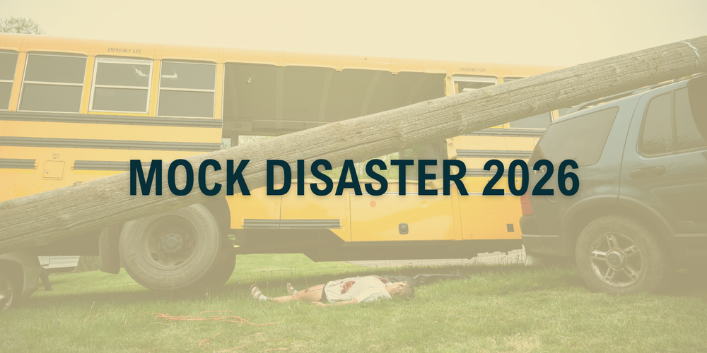 Image of school bus with middle cut out, with a tree pole crashed over a car with an actor laying on the floor in front of it with fake blood on her stomach to simulate a mock disaster. with a yellow overlay that is transparent with the text mock disaster 2026 over it.