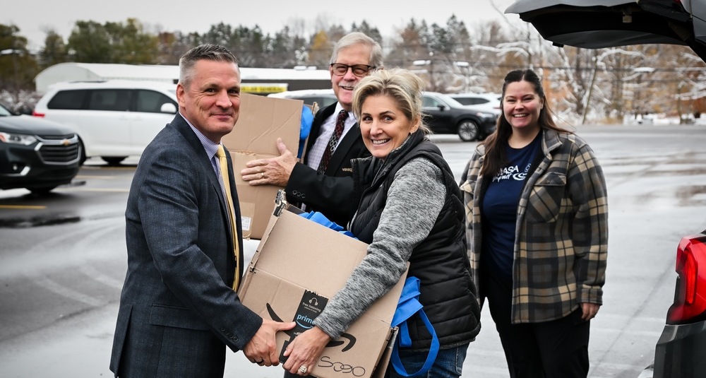 photo of Dave Manson and a woman in jeans and a black vest handing each other a box. with 2 individuals blurred in the background
