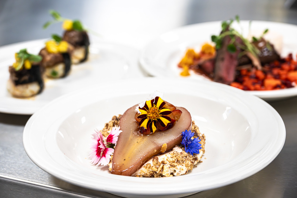 Plated gourmet dish featuring a poached pear slice on a bed of cream and granola, garnished with colorful edible flowers, with two blurred savory dishes in the background on a stainless steel counter.