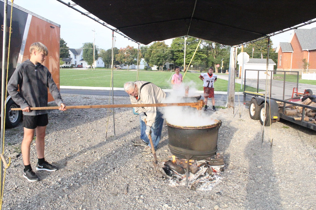 A picture of students continuing to stir the apple butter while Mr. Camper stokes the fire.