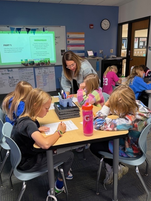 A teacher helping students write down what they are learning on papers at their desks.