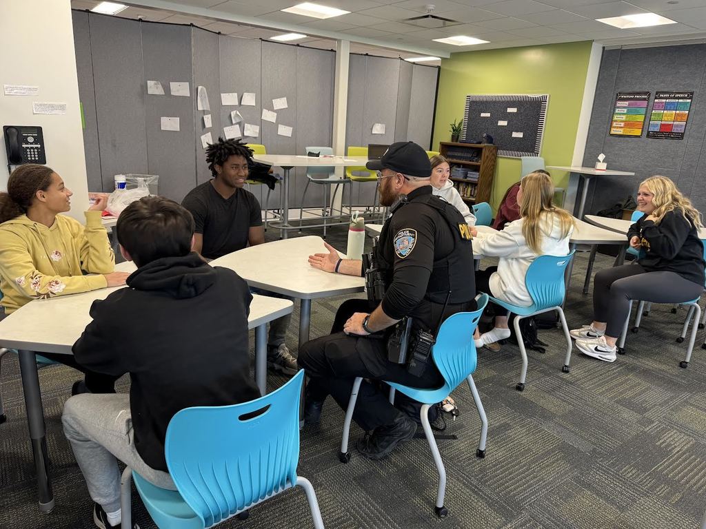 Students sitting at tables across from each other debating. Officer Cooper is debating a student as well.