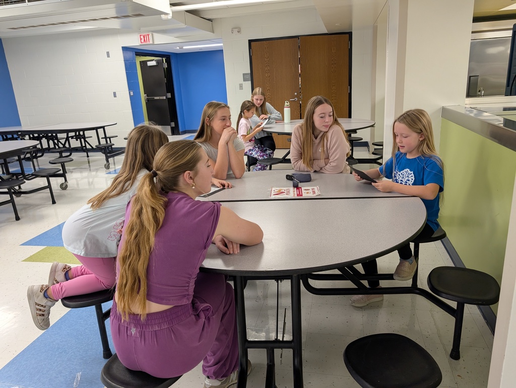 Students sitting at a table listening to one student read to the group.