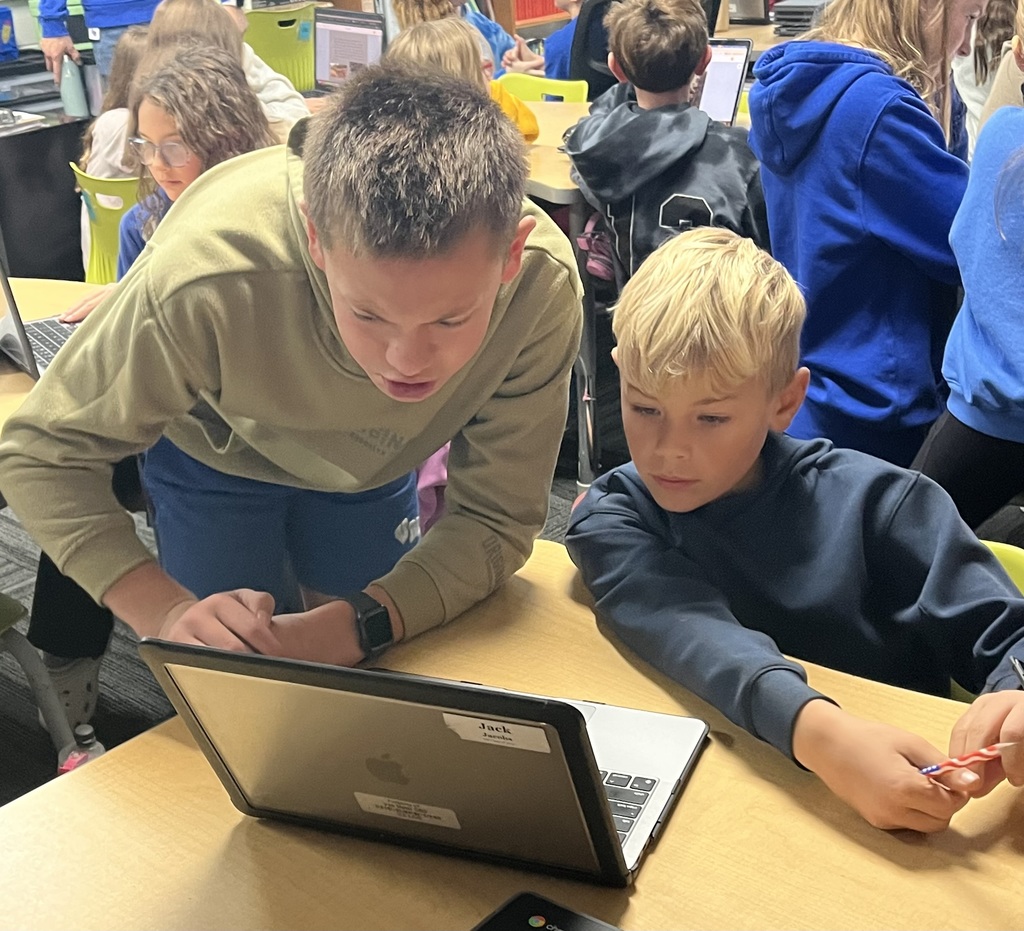 A 7th grader standing at a desk and sharing his ebook with a 3rd grader.