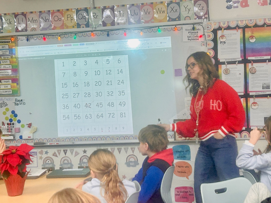 A teacher in a red sweater at the front of the room with the whiteboard displaying numbers in boxes