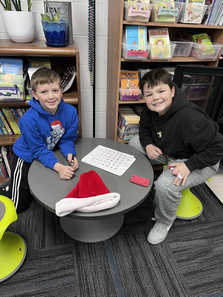 Two children sitting at a small round table in a classroom, working on a worksheet with a Santa hat placed on the table.