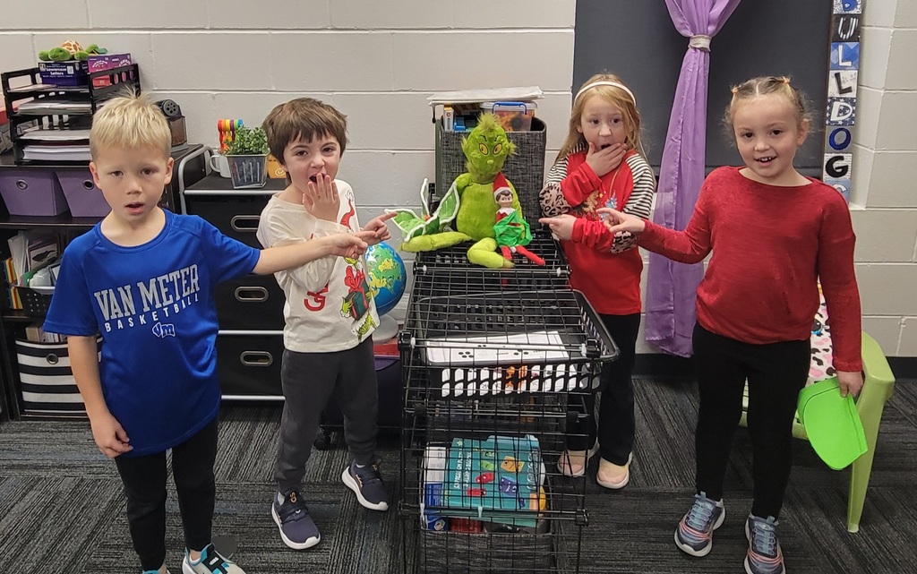 Four children stand around a black wire stand filled with books and a grinch toy with an elf in a classroom setting.