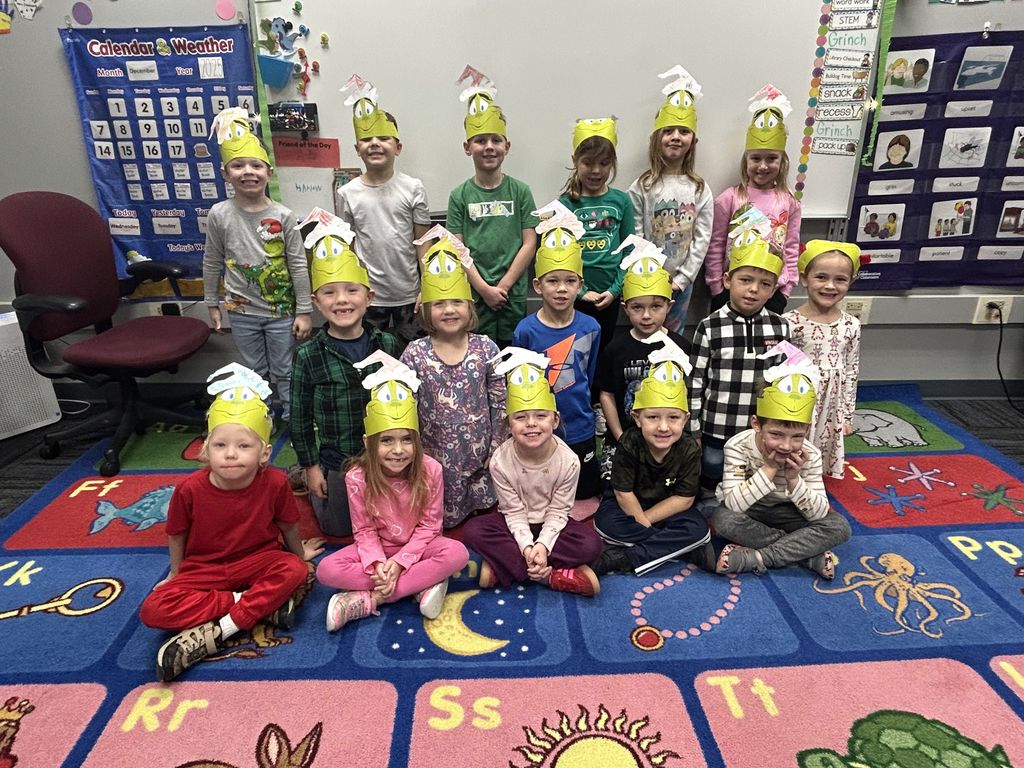 Group of young children wearing handmade grinch hats posing in a colorful classroom with educational posters and alphabet carpet.