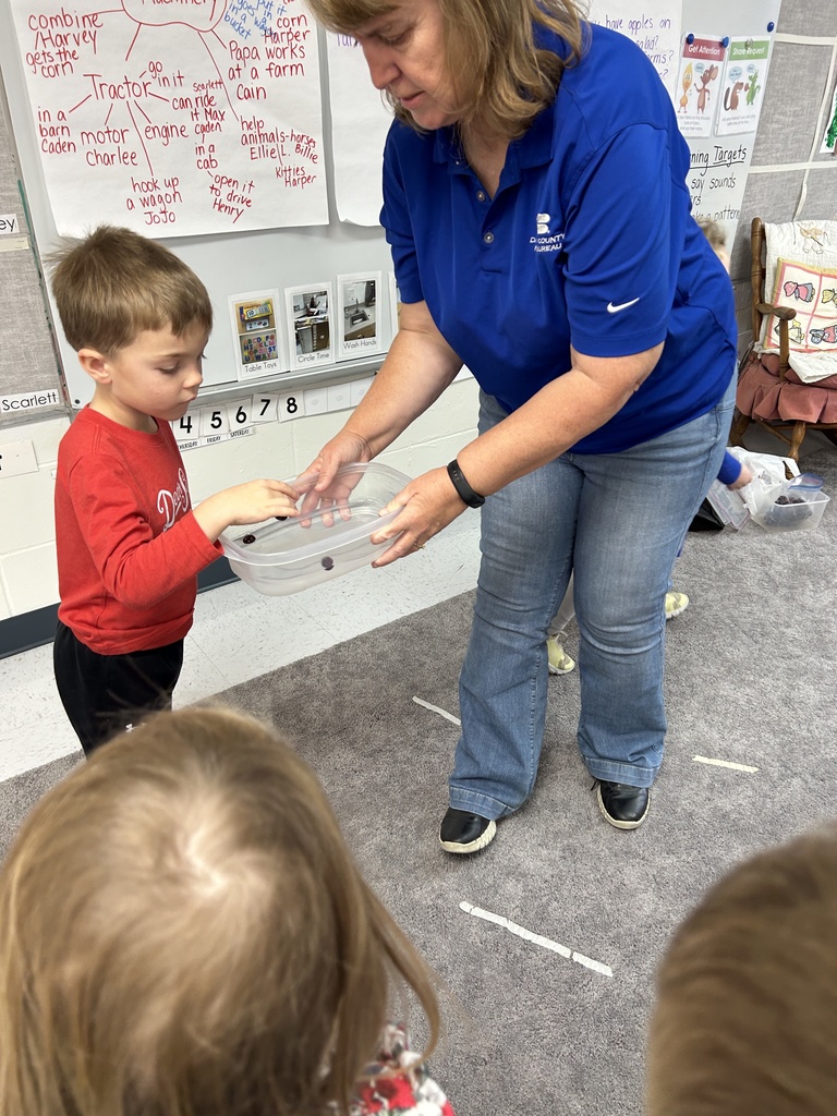 Teacher in blue shirt holding a clear plastic container with a cranberry in it for young boy in a red shirt inside a classroom.