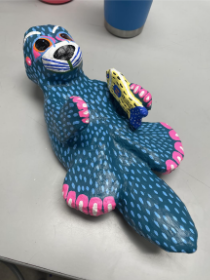 Colorful handcrafted otter figurine holding a yellow fish, painted with blue and pink patterns, resting on a light surface.