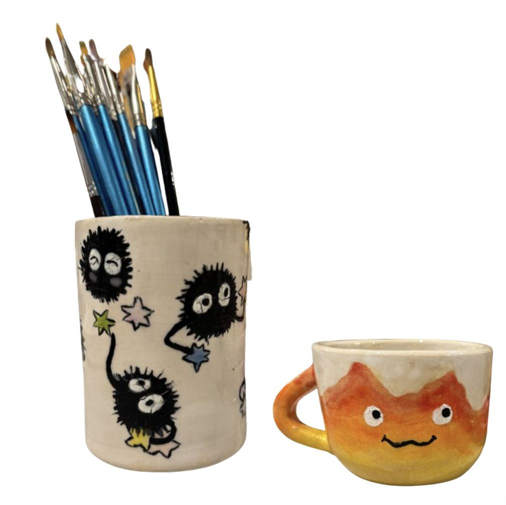 Ceramic cup decorated with black soot sprites holding stars, filled with paintbrushes, next to a small orange-yellow cup with a smiling face and a handle.