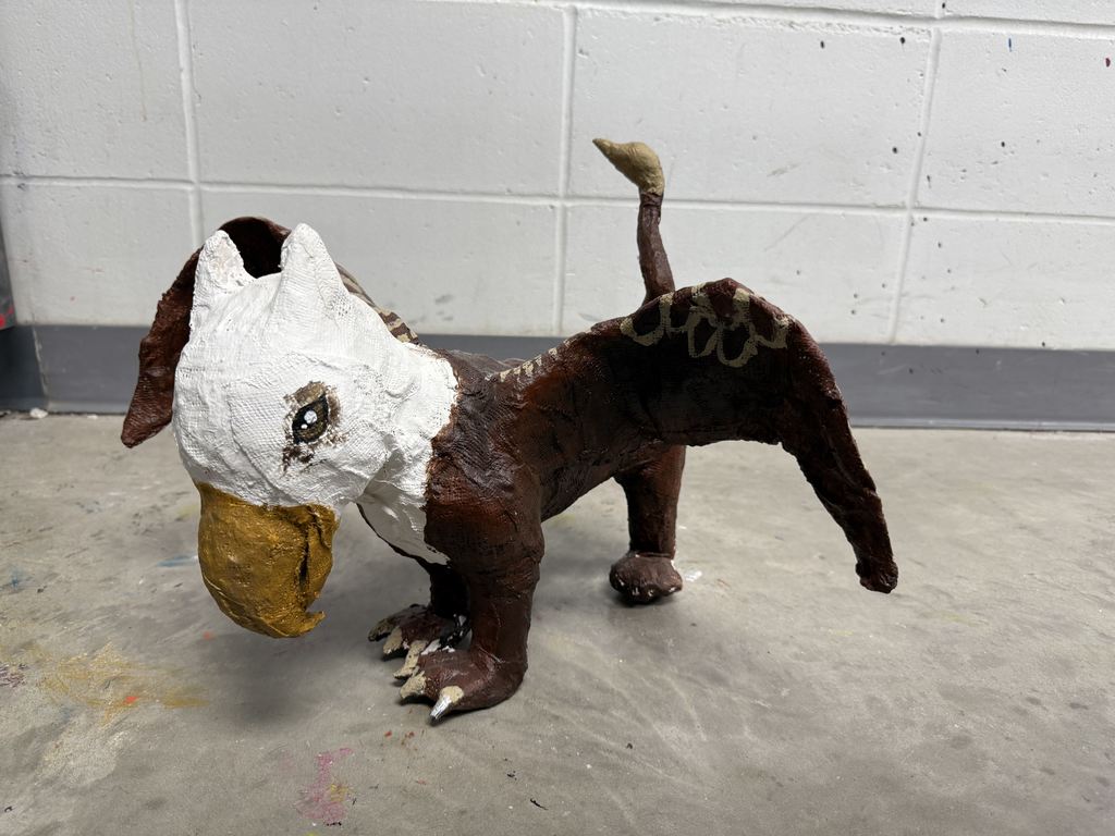 Sculpture of a small animal with a white head, brown body, and a yellow beak standing on a concrete floor.