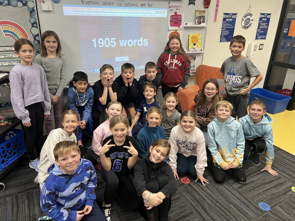 Group of elementary students posing in a classroom with a screen showing '1905 words' behind them.