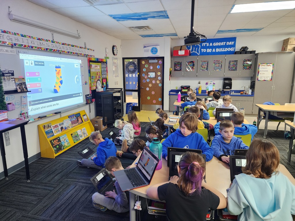 Third grade students in a classroom using laptops and tablets while a coding lesson is projected on the whiteboard.