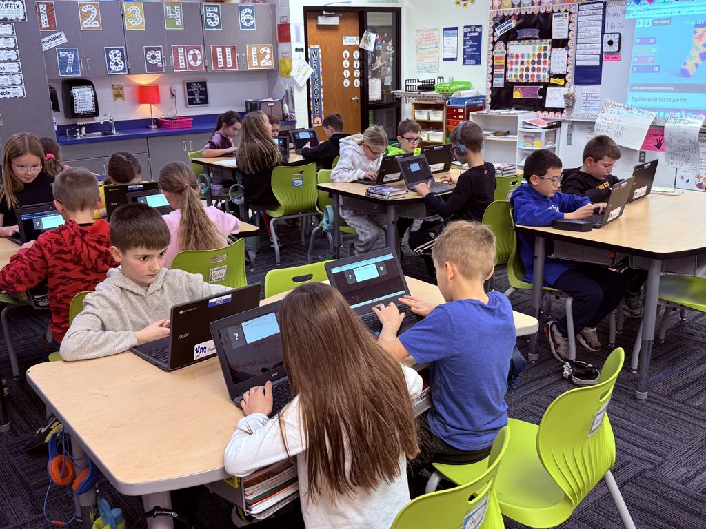 Third grade students focused on laptops in a colorful classroom filled with educational posters and materials.
