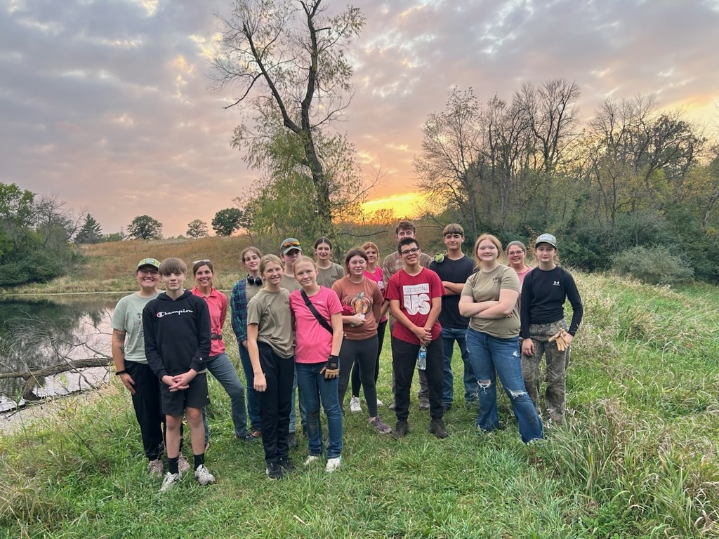 Van Meter's Conservation group pictured outside at sunrise by a body of water, trees, and green plants.