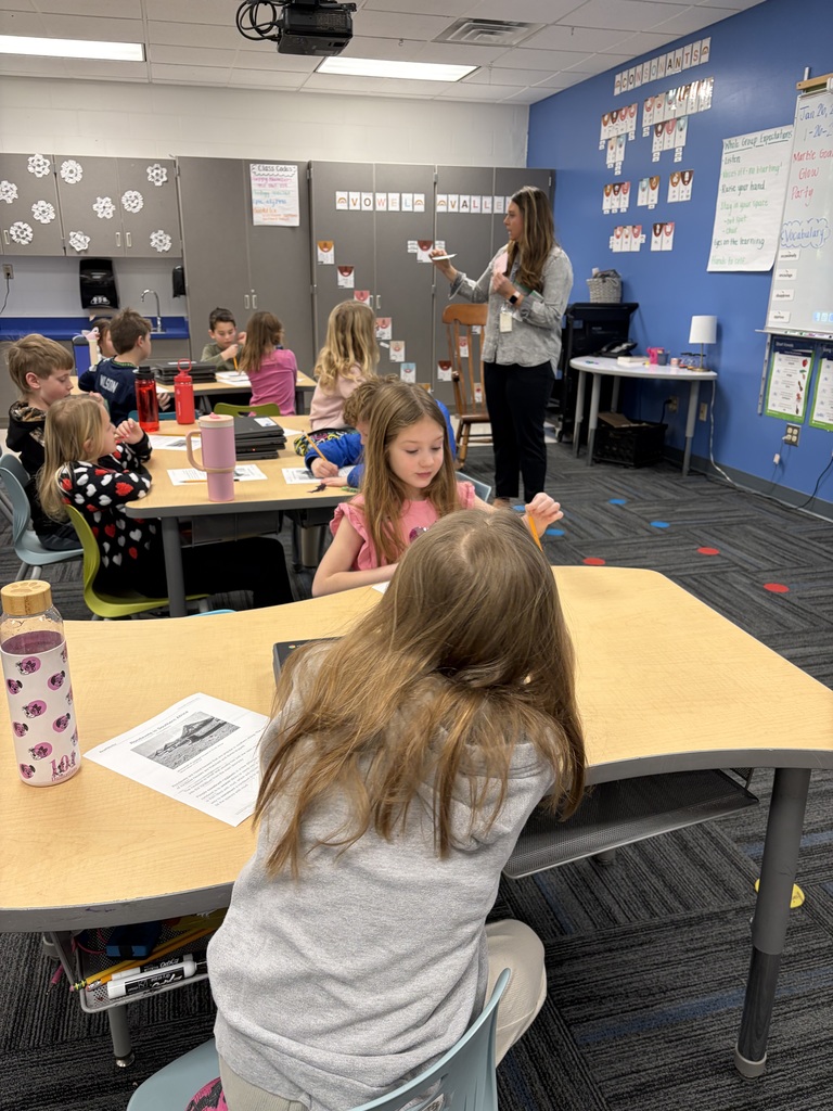 Teacher engaging 2nd grade students in a classroom with colorful educational posters on the walls.