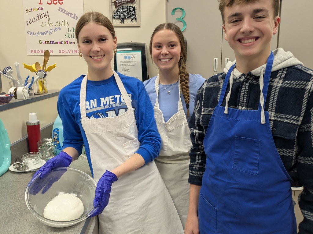 Three Van Meter students wearing aprons and gloves stand in a kitchen classroom, one holding a bowl with dough inside.