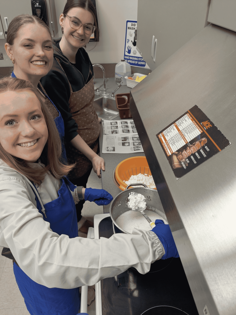 Three Van Meter students wearing aprons and gloves prepare ingredients in a kitchen, focusing on mixing a white substance in a pot on the stove.