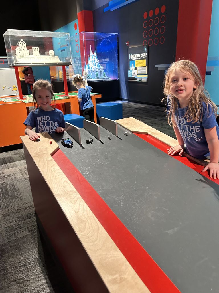 Two VM students playing with small toy cars on a black and red racing table in a brightly lit museum exhibit space.