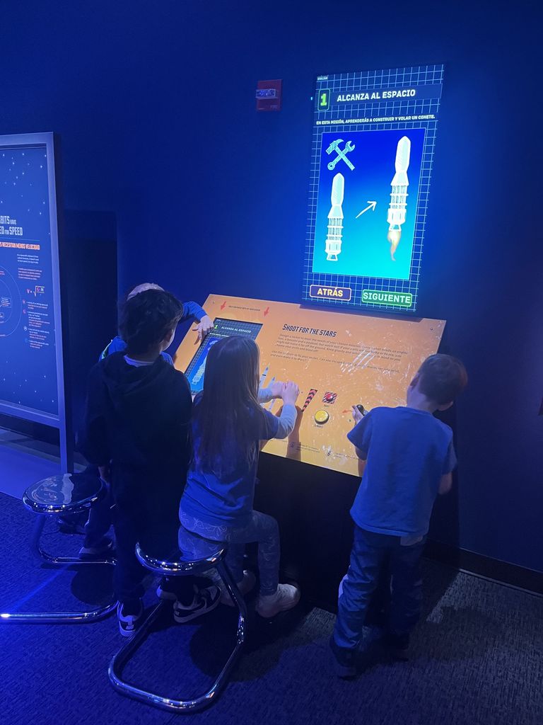 Three children interact with a space-themed educational exhibit featuring a touchscreen and a glowing display about rocket parts in Spanish.