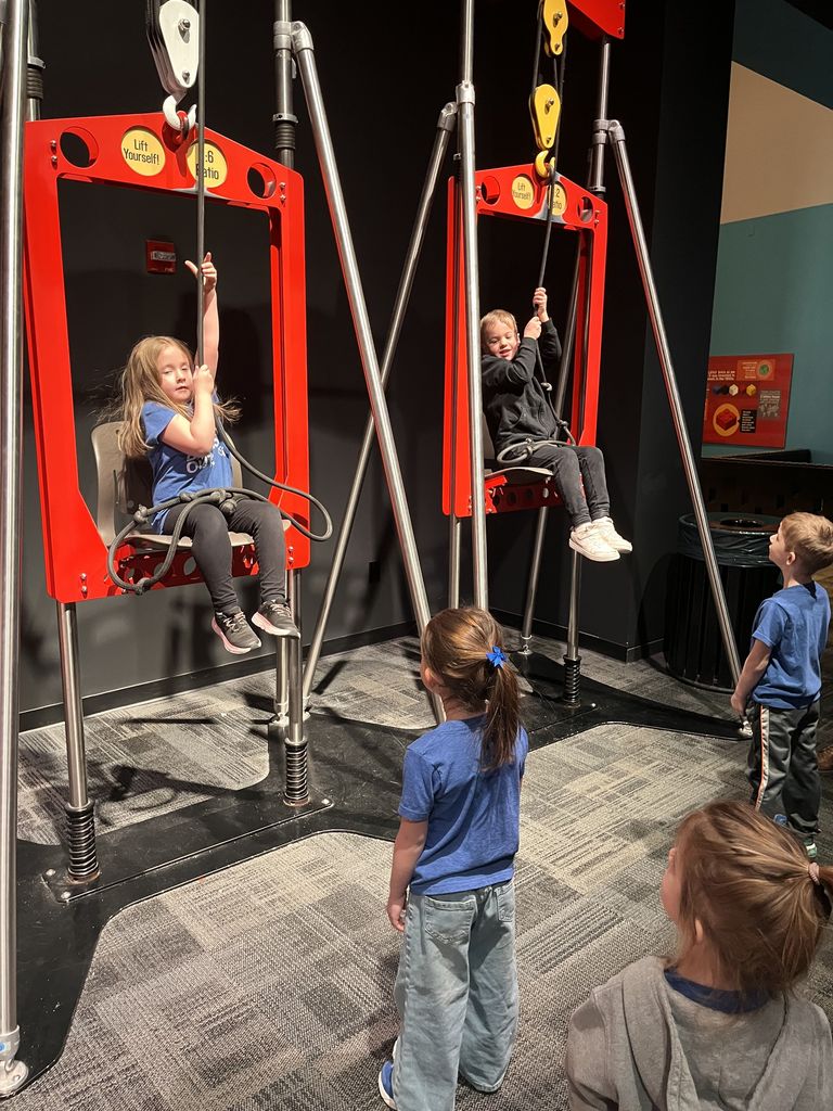 Two children seated on red pulley swings, pulling ropes to lift themselves while three other kids watch nearby in an indoor play area.