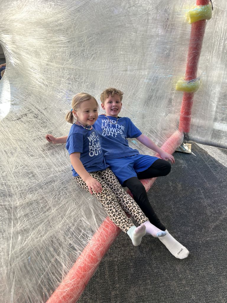 Two VM students sitting side by side on a large, netted play structure with sunlight casting shadows around them.
