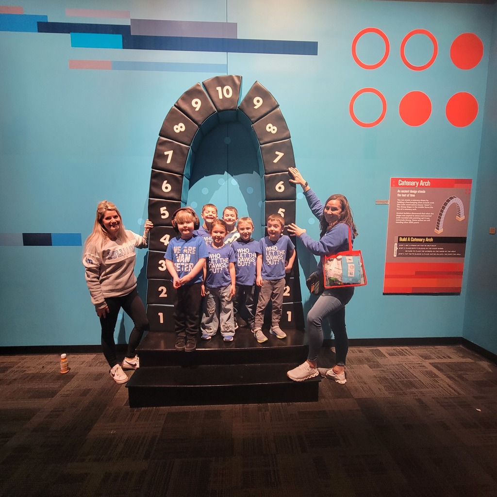 Group of children and two adults posing inside a numbered arch exhibit in a museum setting with blue walls and informational signage.