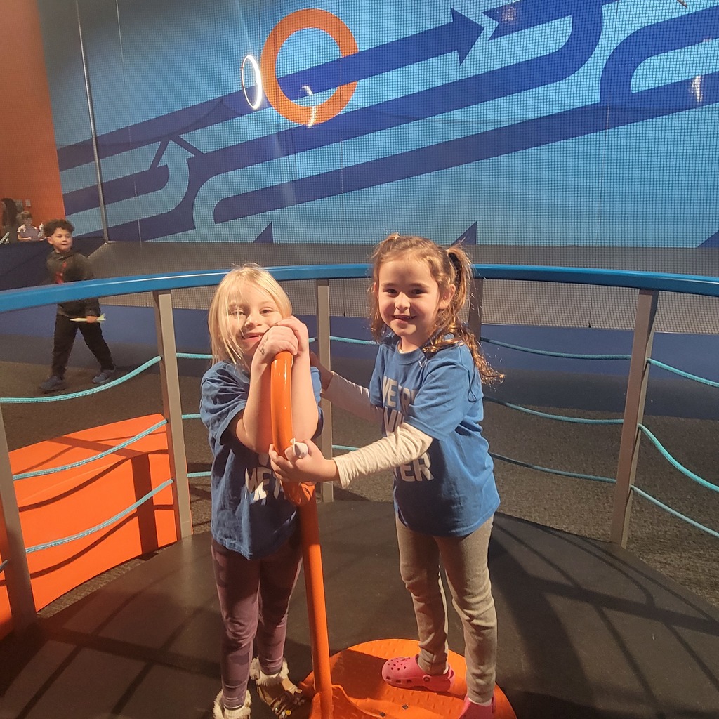 Two young girls in blue shirts play together on an orange and blue interactive exhibit inside a modern museum space.