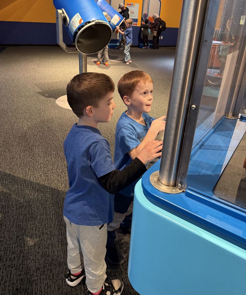 Two young boys in blue shirts interact with a hands-on exhibit at a science museum, exploring its features closely.