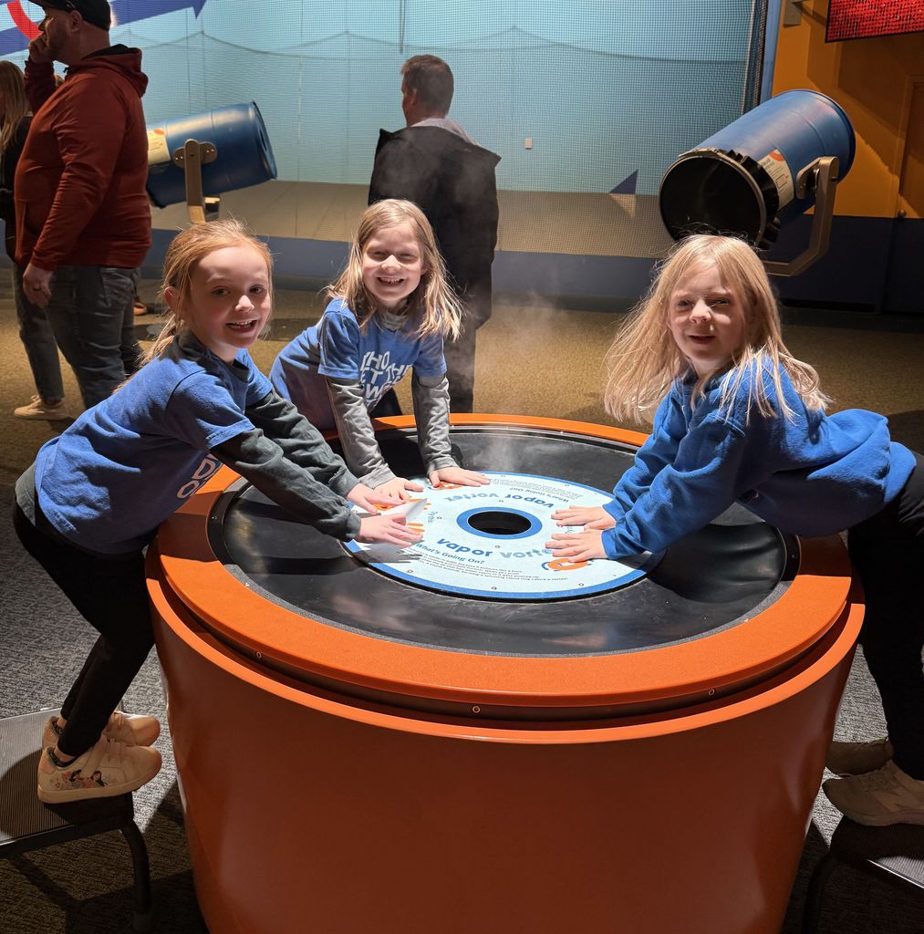 Three children in blue shirts interact with a large orange and black circular exhibit table indoors.