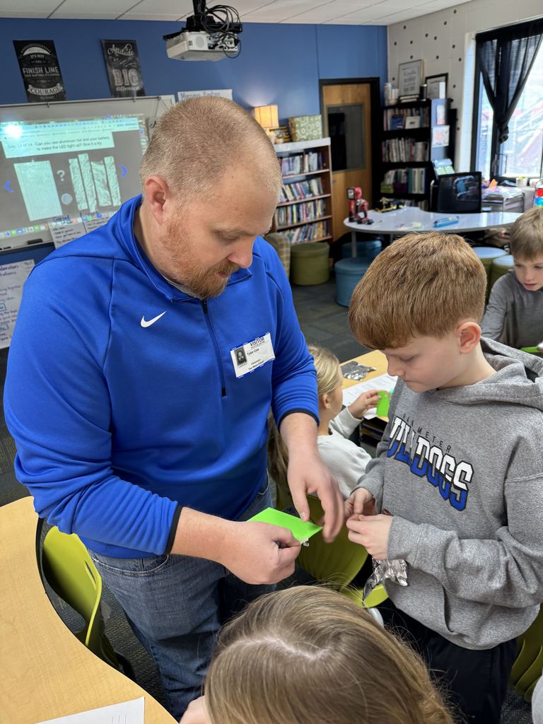 Guest speaker in blue sweatshirt helping young student with a classroom activity involving green paper pieces.