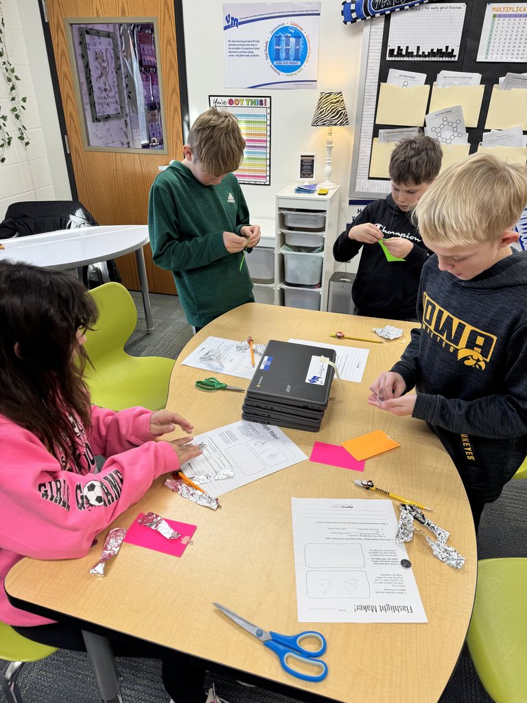 Four children engaged in a classroom activity involving paper folding and crafting at a round table with worksheets and scissors.