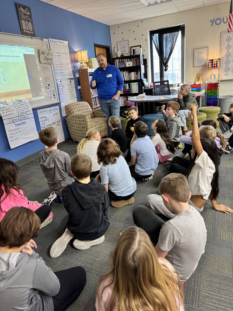 Guest speaker standing near a whiteboard explaining a lesson to attentive elementary students seated on the classroom floor.