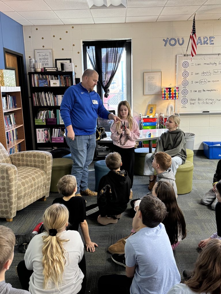 Guest speaker standing and interacting with a student in a classroom while other children sit on the floor watching attentively.