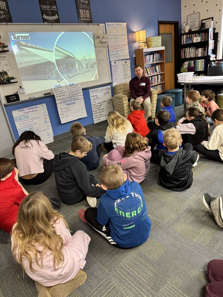 Guest speaker sitting in a classroom reading to a group of attentive young students seated on the floor.