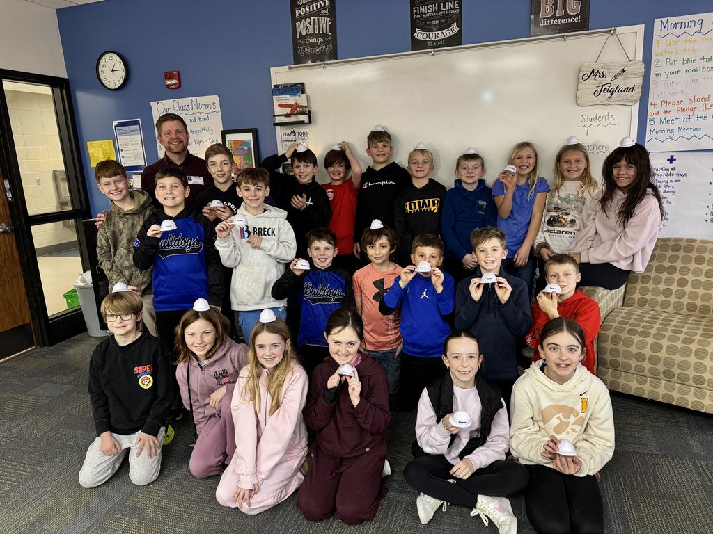 Group of elementary students posing together in a classroom with a whiteboard and motivational posters behind them.