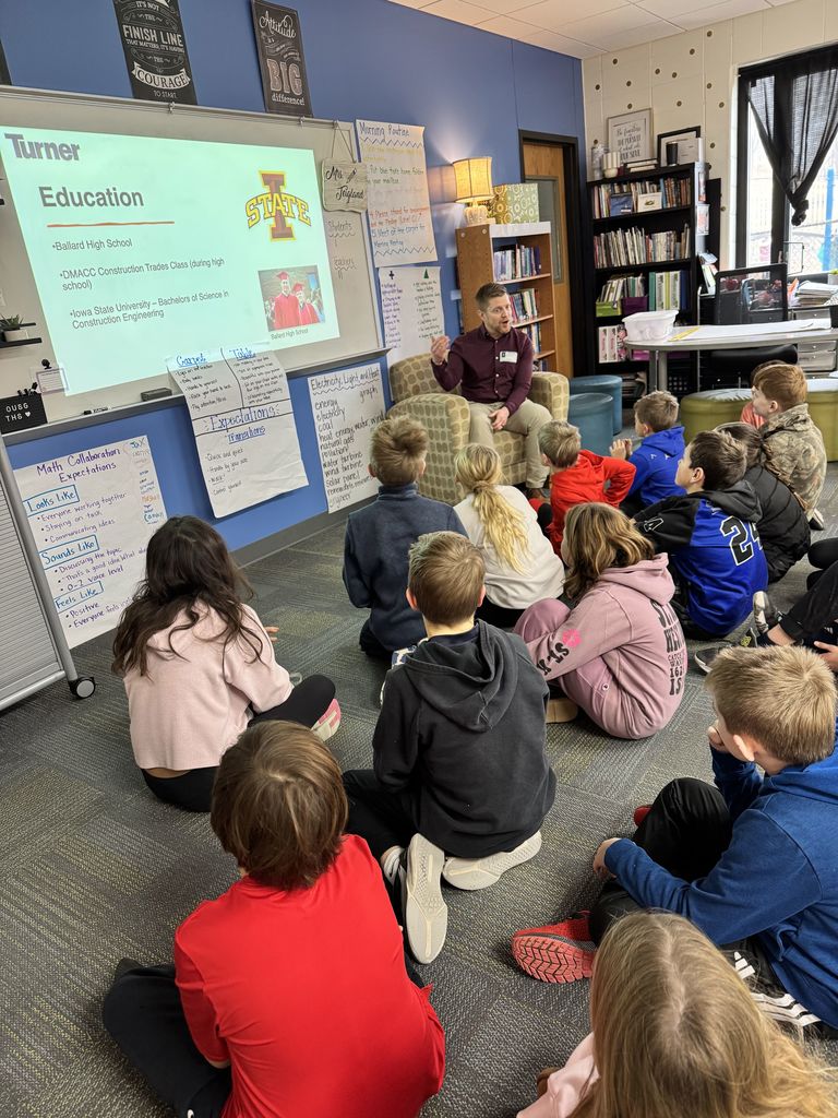 Guest speaker sitting in a classroom reading to a group of attentive young students seated on the floor.