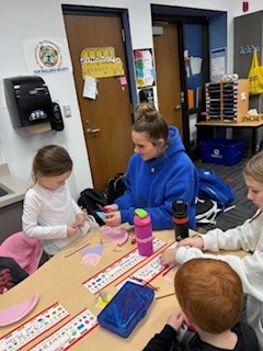 Three students seated at a classroom table engaged in a craft activity with paper and scissors, surrounded by school supplies and colorful water bottles.