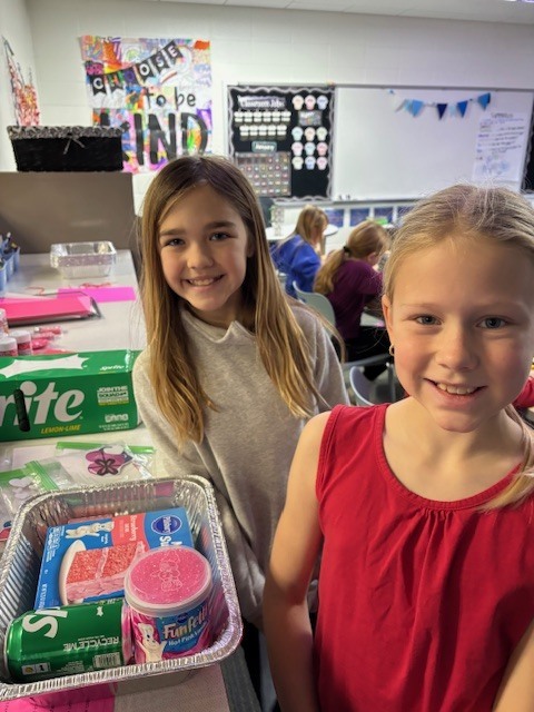 Two young girls stand by a table with baking supplies including frosting, sprinkles, and cake mix in a classroom setting.