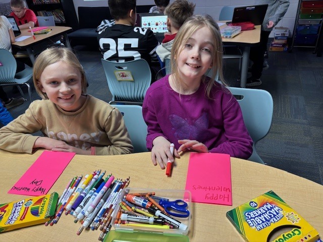 Two students working together at a classroom table with pink paper, markers, and a laptop open in front of them.
