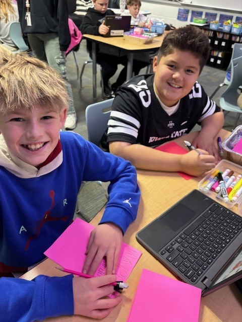 Two students working together at a classroom table with pink paper, markers, and a laptop open in front of them.