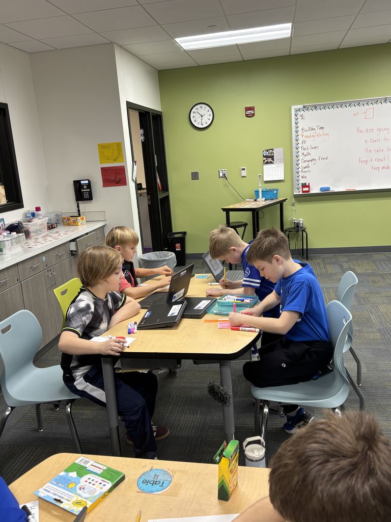 Four young students working on laptops and writing at a classroom table with school supplies scattered around.