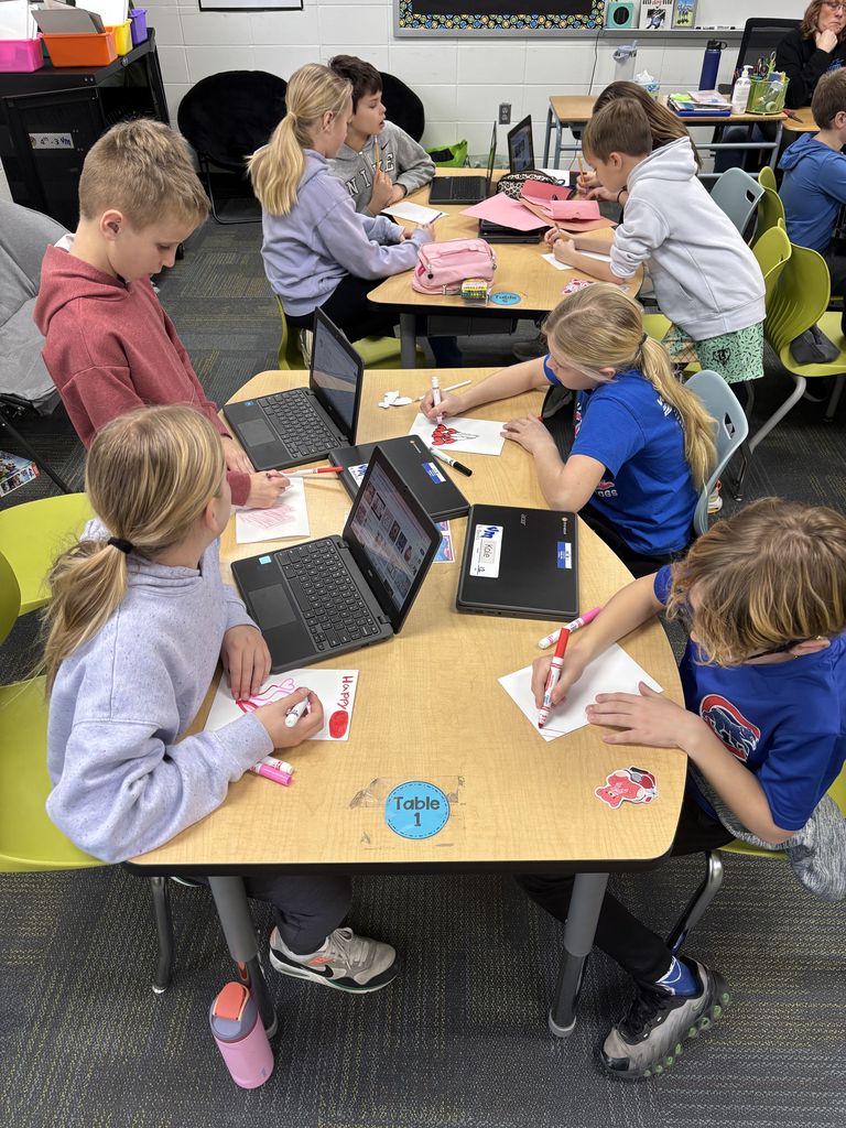 Students working on valentines day cards and writing at a classroom table with school supplies scattered around.