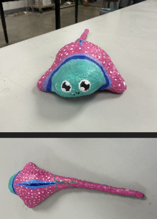 Hand-painted rock shaped like a pink and turquoise stingray with big eyes, displayed on a white table.