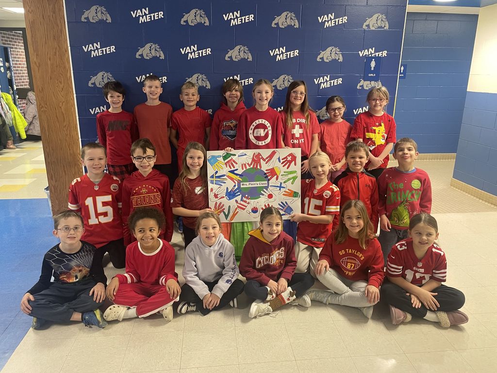 Group of children and two adults wearing red shirts posing with a colorful handprint poster in front of a Van Meter backdrop.