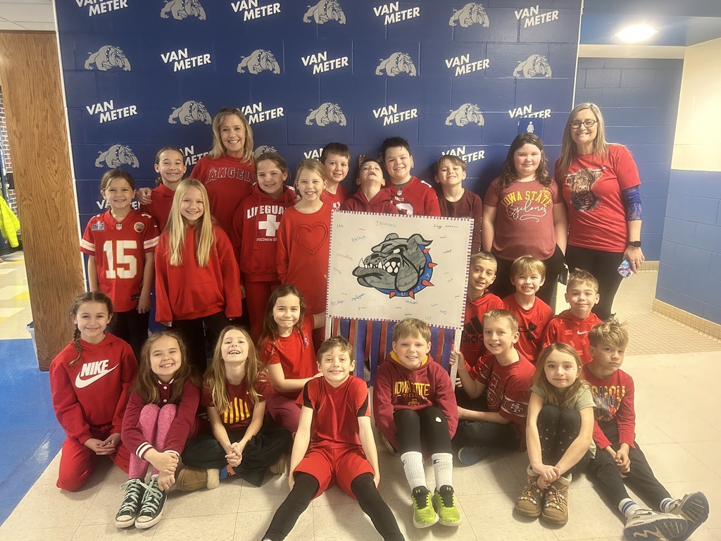 Group of children and adults wearing red and maroon sportswear posing indoors with a bulldog mascot poster in front of a Van Meter backdrop.